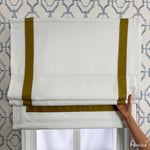 Close-up of a hand adjusting flat Roman shades in white with mustard yellow trim. Customizable window treatment with easy installation.